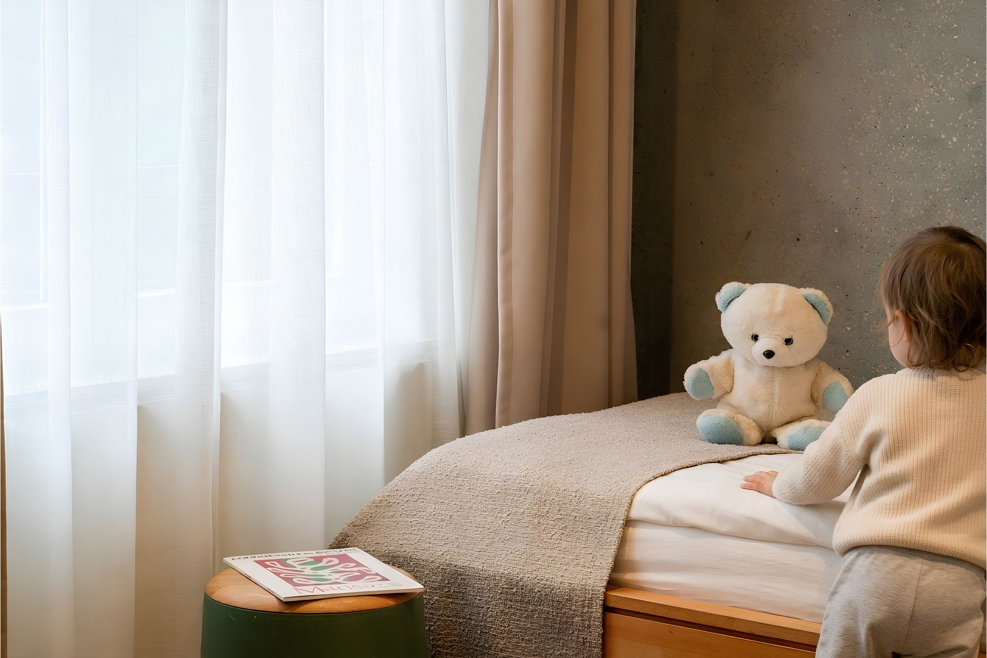 Photographe d’hôtel en Alsace : enfant et ours en peluche sur un lit, chambre aux rideaux clairs