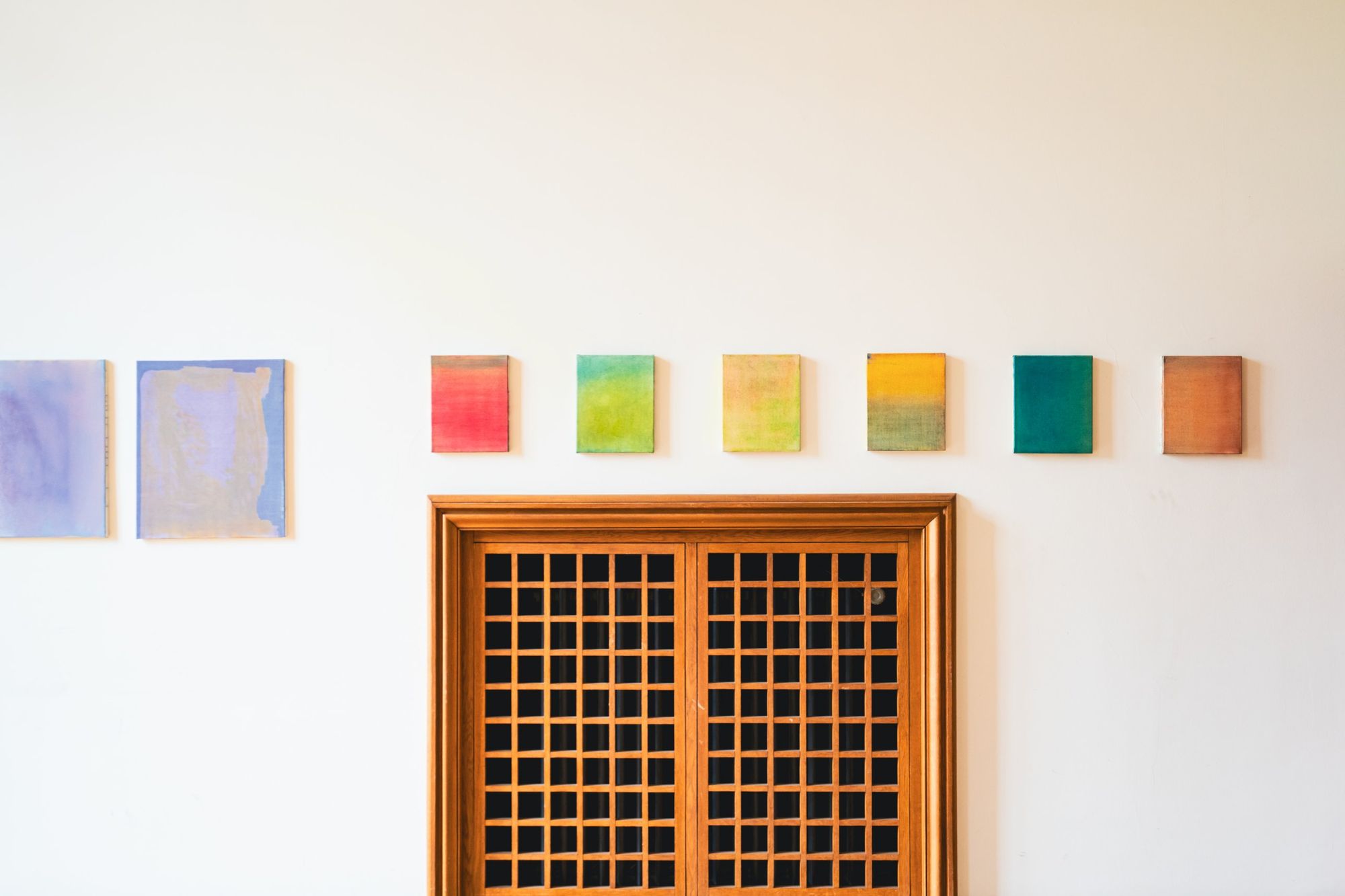 Photographe d’exposition à Strasbourg : petites toiles colorées alignées sur un mur blanc au-dessus d’une porte en bois à croisillons