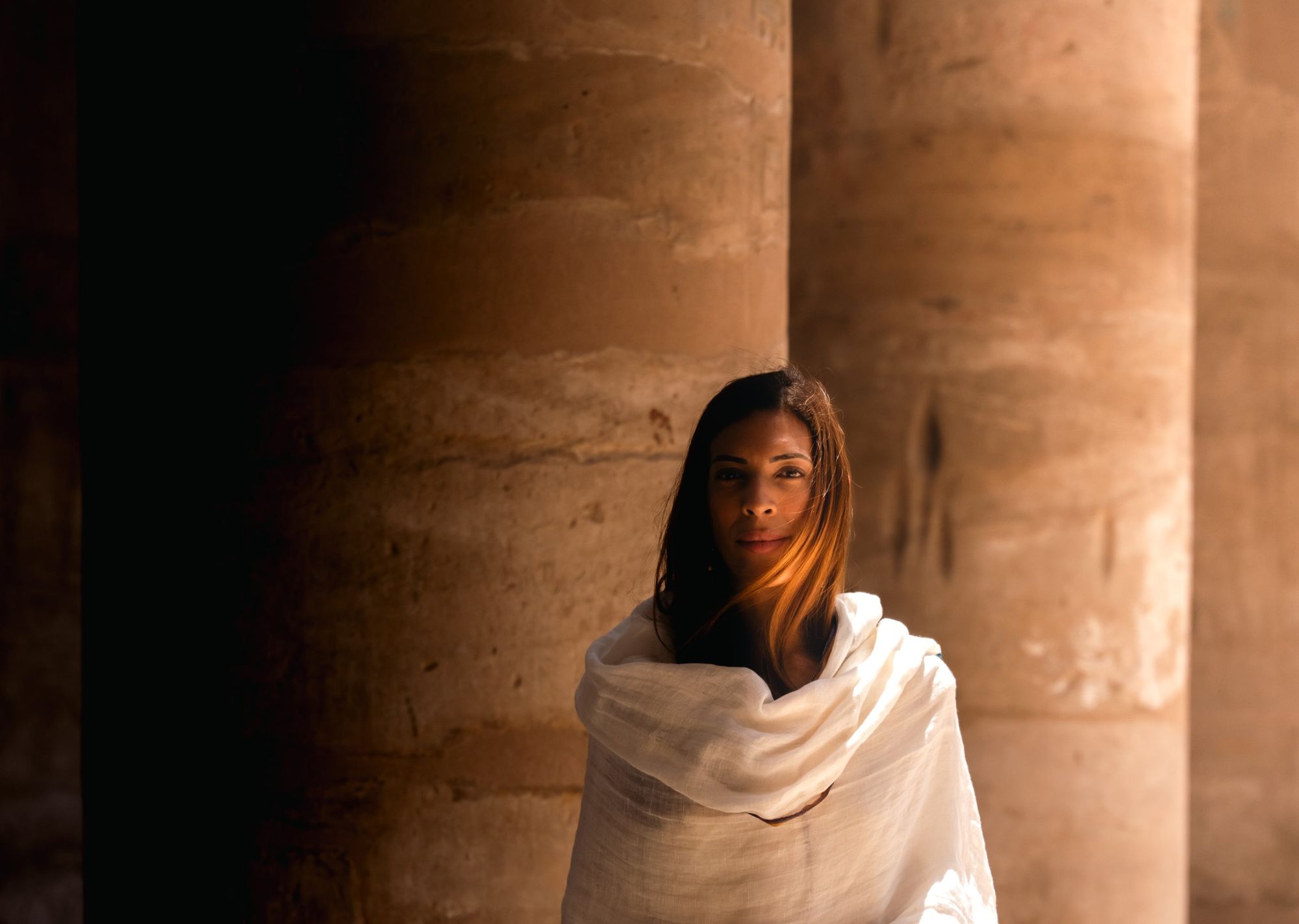 Shooting photo : portrait d’une femme drapée dans un châle blanc entre les colonnes de pierre d’un temple, lumière chaude