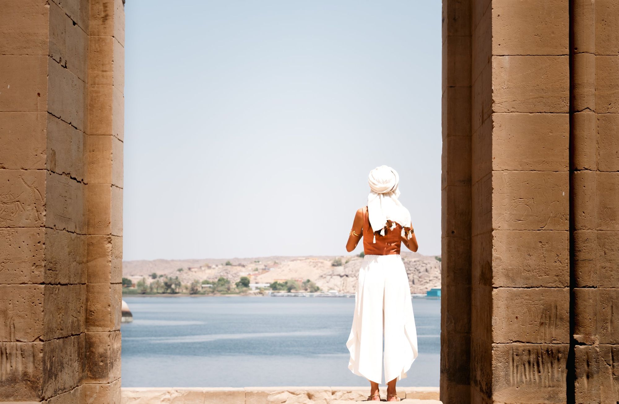 Shooting photo en voyage : femme en tenue blanche et turban de dos, cadrée entre deux piliers de pierre face au lac